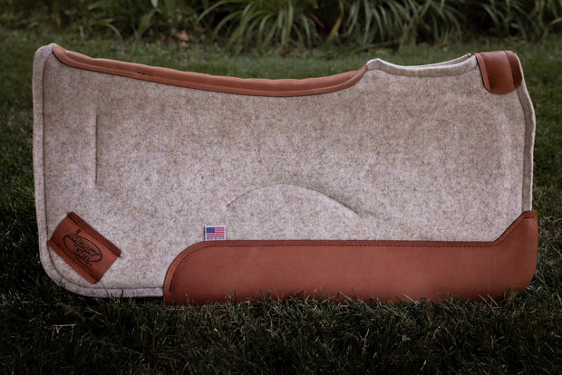 Contoured saddle pad in tan felt with burnt orange colored leather  standing up in a field of grass